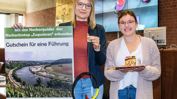 Eine blonde Frau mit Brille und eine junge Frau halten ein Schild mit Informationen zu den Neckarguides und einem Gutschein für eine Führung. Im Hintergrund ist ein Bildschirm mit bunten Objekten zu sehen.