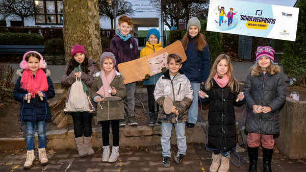 Eine Gruppe von zehn Kindern steht im Freien vor einem Baum. Sie halten verschiedene Gegenstände in den Händen und lächeln in die Kamera. Im Hintergrund ist ein Gebäude zu sehen. Oben rechts befindet sich ein Banner mit dem Text „Schulwegprofi“.