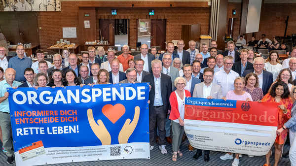 Die Abbildung zeigt eine Gruppe von über 50 Personen in einem Versammlungsraum, die gemeinsam Plakate zur Organspende halten. Die Plakate weisen auf die Wichtigkeit der Organspende hin und fordern dazu auf, sich zu informieren und Entscheidungen zu treffen, um Leben zu retten.