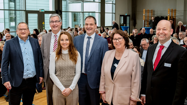 Sechs Personen stehen zusammen in einem großen Raum mit vielen sitzenden Menschen im Hintergrund. Sie lächeln freundlich in die Kamera. Die Szene vermittelt eine positive Atmosphäre, möglicherweise bei einer Veranstaltung oder Versammlung.