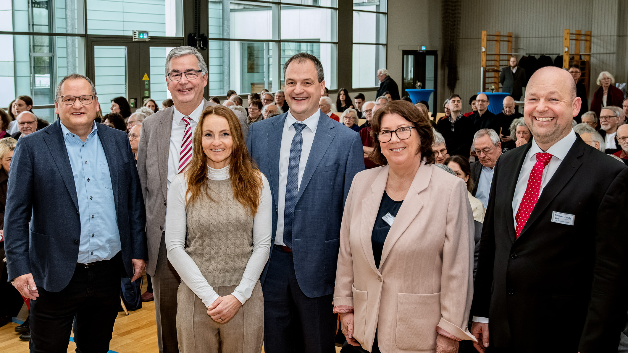 Sechs Personen stehen zusammen in einem großen Raum mit vielen sitzenden Menschen im Hintergrund. Sie lächeln freundlich in die Kamera. Die Szene vermittelt eine positive Atmosphäre, möglicherweise bei einer Veranstaltung oder Versammlung.