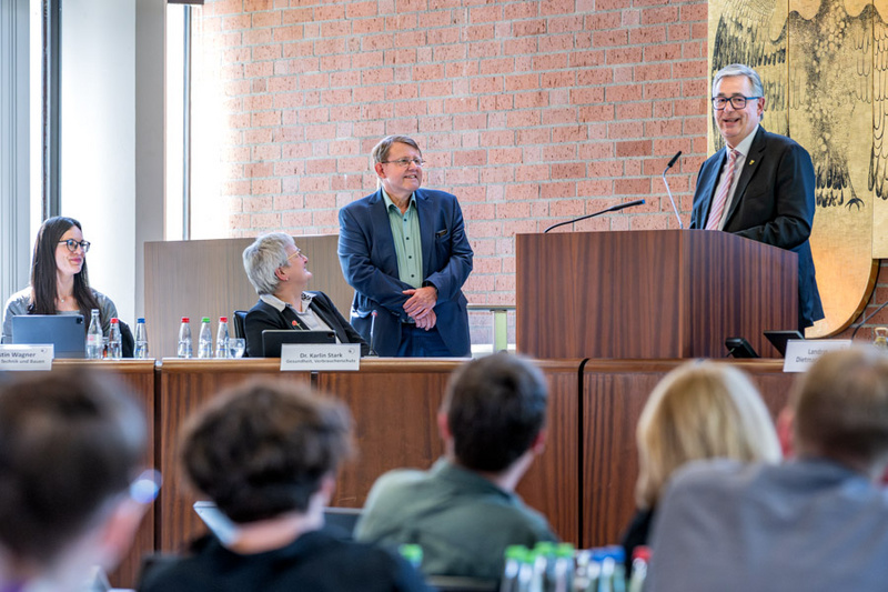 Eine Podiumsdiskussion findet statt, an der mehrere Personen teilnehmen. Ein Mann steht an einem Rednerpult und spricht, während die anderen Teilnehmer ihm aufmerksam zuhören. Im Hintergrund ist eine Etwand mit einer künstlerischen Darstellung zu sehen.