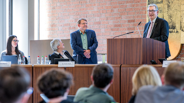 Eine Podiumsdiskussion findet statt, an der mehrere Personen teilnehmen. Ein Mann steht an einem Rednerpult und spricht, während die anderen Teilnehmer ihm aufmerksam zuhören. Im Hintergrund ist eine Etwand mit einer künstlerischen Darstellung zu sehen.