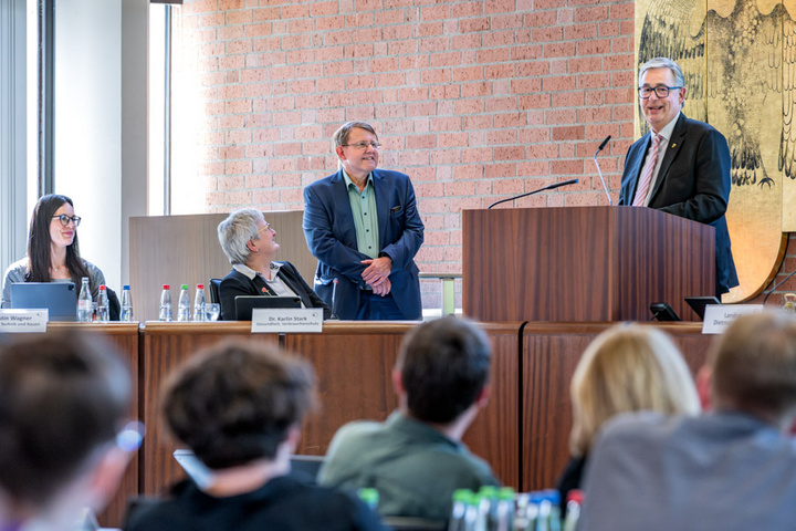 Eine Podiumsdiskussion findet statt, an der mehrere Personen teilnehmen. Ein Mann steht an einem Rednerpult und spricht, während die anderen Teilnehmer ihm aufmerksam zuhören. Im Hintergrund ist eine Etwand mit einer künstlerischen Darstellung zu sehen.