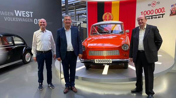 Three men stand in front of an orange Trabant car, marked with the number "1000000." Behind them is a backdrop featuring the German flag. The setting appears to be a modern exhibition space celebrating the car's significance.