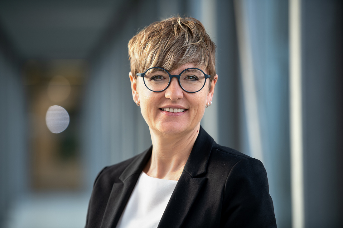 Stefanie Bartzsch Eine Frau mit kurzen, blonden Haaren und einer modernen Brille lächelt freundlich. Sie trägt ein schwarzes Blazer und ein weißes Oberteil. Der Hintergrund zeigt einen hellen, verschwommenen Gang mit Fensterlicht.