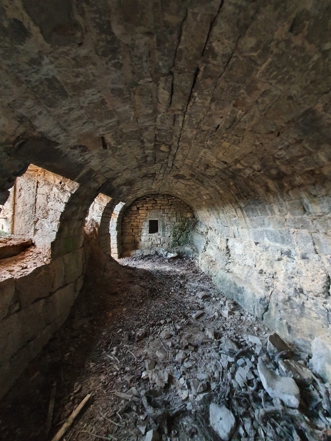 Ein verfallener, gewölbter Raum mit Steinwänden und einer unebenen, mit Schutt bedeckten Bodenfläche. An einer Wand ist ein kleines Fenster sichtbar, und das Licht fällt in den Raum, wobei die Struktur und der Verfall der Umgebung betont werden.