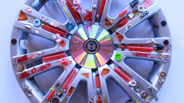 Una ruota decorativa di metallo, con un centro colorato e varie applicazioni di adesivi, colori e piccole decorazioni. La superficie presenta un mix vivace di tonalità e forme, dando un aspetto artistico e creativo all'oggetto.