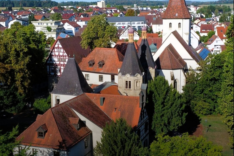 Luftaufnahme eines historischen Gebäudes in Hemmingen, umgeben von Bäumen und Wohnhäusern. Im Vordergrund sind ein Rathaus und ein Kirchturm sichtbar. Darüber steht der Text „Hemmingen ENTDECKE UNSERE REGION“. (Text KI generiert)