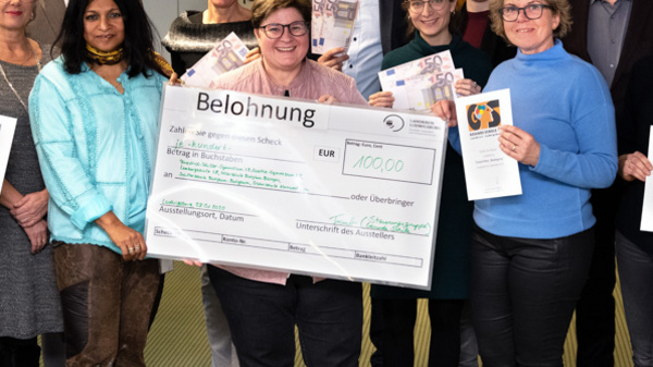 A diverse group of twelve people stands together in an office setting, holding a large check for 1,000 euros and various documents. The individuals are smiling, celebrating a recognition or award, with certificates and promotional materials visible in their hands.