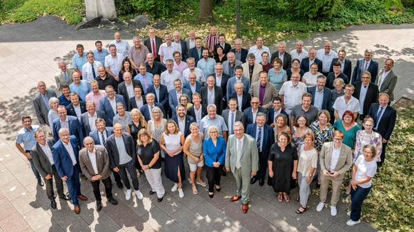 Eine große Gruppe von über 60 Personen steht im Freien, umgeben von Bäumen und Grünflächen. Die Menschen sind in formeller und lässiger Kleidung, verteilt auf mehreren Ebenen. Sie lächeln und posieren gemeinsam für ein Gruppenfoto.
