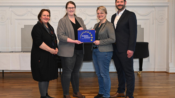 Vier Personen stehen zusammen in einem Raum mit Parkettboden. Sie halten gemeinsam ein Schild mit dem Aufdruck "Europa" und den Sternen der Europäischen Union. Die Frauen tragen formelle Kleidung, während der Mann einen Anzug trägt. Der Hintergrund zeigt eine elegante Wand mit Dekorationen.