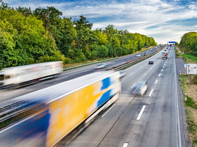 Eine vielbefahrene Autobahn mit mehreren Fahrspuren. Verschiedene Lkw und Autos bewegen sich schnell in beide Richtungen. Im Hintergrund ist ein bewaldeter Bereich zu sehen, der die Straße umgibt. Der Himmel ist teilweise bewölkt.