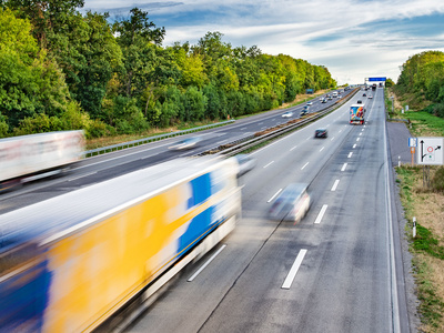 Eine vielbefahrene Autobahn mit mehreren Fahrspuren. Verschiedene Lkw und Autos bewegen sich schnell in beide Richtungen. Im Hintergrund ist ein bewaldeter Bereich zu sehen, der die Straße umgibt. Der Himmel ist teilweise bewölkt.