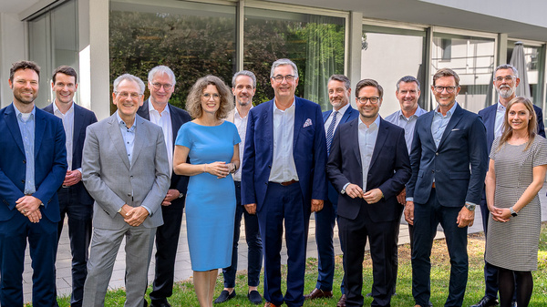 Eine Gruppe von 15 Personen steht in einem Garten vor einem modernen Gebäude. Sie tragen formelle Kleidung und lächeln. In der Mitte der Gruppe steht eine Frau in einem blauen Kleid. Der Hintergrund zeigt einen grünen Rasen und einen hellen Himmel.