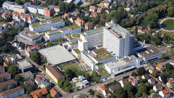 Das Bild zeigt eine luftansicht eines weitläufigen Krankenhausgeländes mit mehreren Gebäuden, darunter ein hoher Hauptbau und angrenzende Flächen mit Parkplätzen. Umgeben ist das Gelände von Wohngebieten und Bäumen.