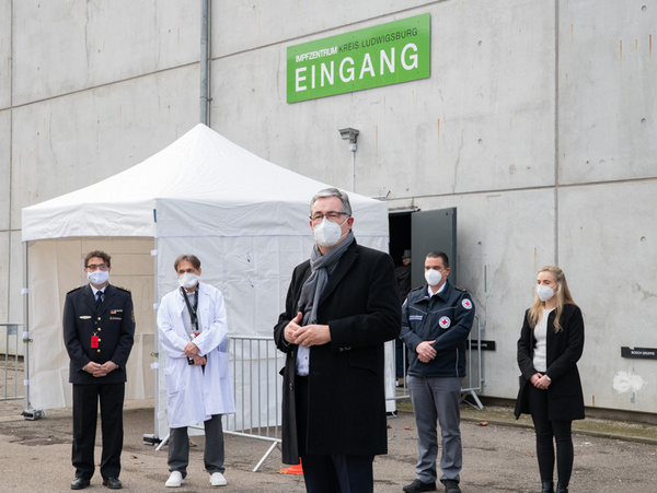 14.10.2021 - Eröffnung des Kreisimpfzentrums in Ludwigsburg Ein Mann mit Brille und Mundschutz steht vor einem Eingang mit dem Schild „EINGANG“. Hinter ihm stehen mehrere Personen, darunter ein Arzt und Mitarbeiter des Roten Kreuzes, ebenfalls mit Mundschutz. Im Hintergrund ist ein Zelt zu sehen, das für eine Veranstaltung vorbereitet ist.