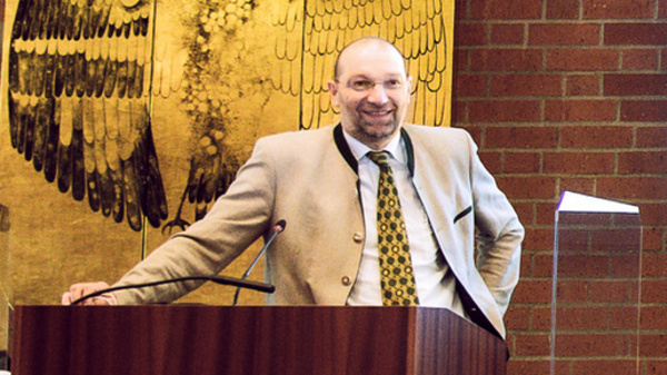 Three speakers stand at a podium in a conference setting. A large screen behind them displays the text "Herzlich Willkommen" (Welcome) and the event title regarding death certificates. The audience area and table setups suggest a formal presentation.