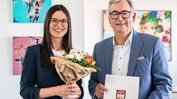 Ein Mann und eine Frau stehen lächelnd in einem modernen Raum. Die Frau hält einen Blumenstrauß, während der Mann ein Dokument mit dem Wappen eines Bundeslandes in der Hand hält. Im Hintergrund sind bunte Kunstwerke an den Wänden zu sehen.