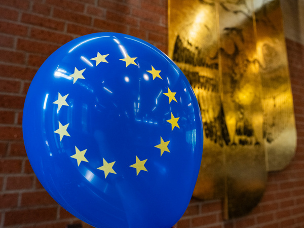 Ein blauer Luftballon mit dem EU-Flaggenmotiv, das zwölf goldene Sterne zeigt, steht im Vordergrund. Im Hintergrund ist ein goldener Adler zu sehen, der symbolisch für Deutschland steht.