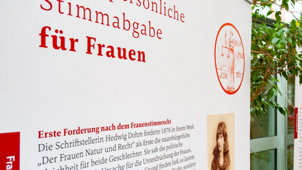 Immagine di una mostra con pannelli informativi che trattano il tema dei diritti di voto per le donne. I pannelli includono testi in tedesco e mostrano fotografie storiche. Sullo sfondo si vedono piante in un ambiente interno.