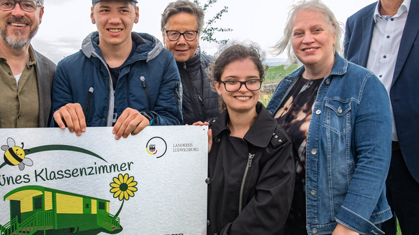 Un gruppo di sei persone sorride davanti a un cartello che rappresenta un'aula scolastica all'aperto chiamata "Grünes Klassenzimmer". Il cartello include disegni di fiori e un codice QR. L'ambiente è verde, con alberi sullo sfondo, e il cielo è nuvoloso.