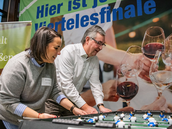 13.02.2024 - After-Work-Kicker-Turnier mit den Mitarbeitenden Zwei Personen spielen Tischfußball. Eine Frau mit dunklem Haar in einem grauen Pullover lächelt, während ein Mann mit Brille daneben steht und konzentriert spielt. Im Hintergrund ist ein Banner mit der Aufschrift "Hier ist das Viertelfinale" und Bildern von Weingläsern zu sehen.