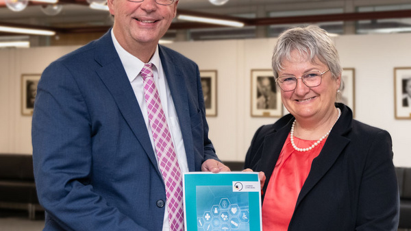 Zwei Personen, ein Mann in Anzug mit Krawatte und eine Frau in Blazer, stehen zusammen und halten einen Bericht mit dem Titel "Gesundheitsbericht". Beide lächeln in die Kamera. Im Hintergrund sind unscharfe Büroumgebungen zu sehen.