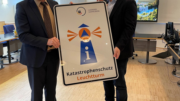Deux hommes se tiennent debout, chacun tenant un panneau représentant un phare, avec le texte "Katastrophenschutz Leuchtturm". Ils semblent participer à un événement officiel. L'image évoque un message sur la protection contre les catastrophes.