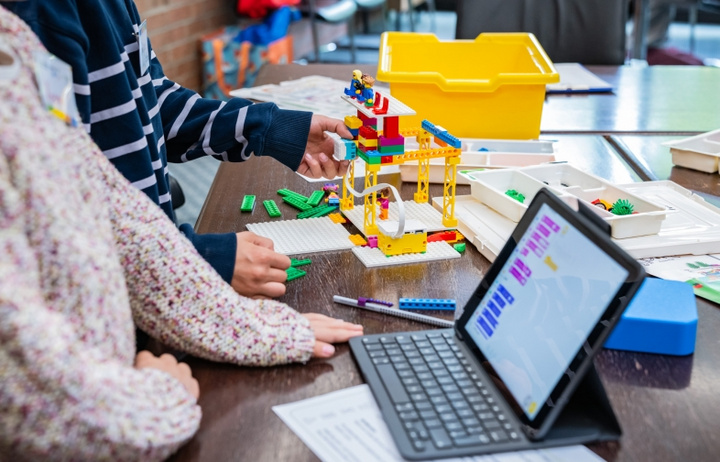Zwei Kinder arbeiten gemeinsam an einem Tisch. Eines von ihnen hält Bausteine, während das andere ein buntes Bauwerk aus Legosteinen konstruiert. Neben ihnen befindet sich ein Tablet, auf dem eine digitale Bauanleitung angezeigt wird. Ein gelbes Behältnis liegt ebenfalls auf dem Tisch.