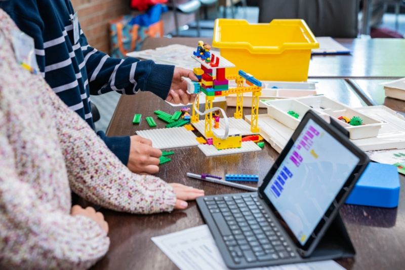 Zwei Kinder arbeiten gemeinsam an einem Tisch. Eines von ihnen hält Bausteine, während das andere ein buntes Bauwerk aus Legosteinen konstruiert. Neben ihnen befindet sich ein Tablet, auf dem eine digitale Bauanleitung angezeigt wird. Ein gelbes Behältnis liegt ebenfalls auf dem Tisch.
