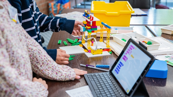 Zwei Kinder arbeiten gemeinsam an einem Tisch. Eines von ihnen hält Bausteine, während das andere ein buntes Bauwerk aus Legosteinen konstruiert. Neben ihnen befindet sich ein Tablet, auf dem eine digitale Bauanleitung angezeigt wird. Ein gelbes Behältnis liegt ebenfalls auf dem Tisch.
