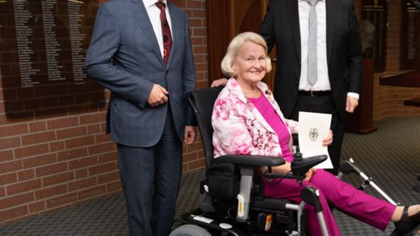 Three individuals are positioned in an indoor setting. A woman in a wheelchair, dressed in pink, smiles as she holds a document. Two men stand beside her, one in a suit and the other in business attire. A floral arrangement decorates the scene, alongside flags in the background.
