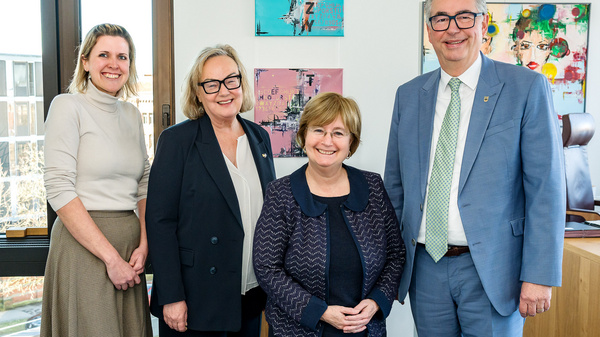 Vier Personen stehen in einem Büro vor einem Fenster. Sie lächeln in die Kamera. Eine Frau trägt einen langen Rock, die anderen sind in Hosen und Blazern gekleidet. Im Hintergrund sind farbige Kunstwerke an der Wand zu sehen.