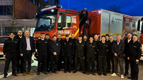 Gruppennfoto von Feuerwehrleuten vor einem Feuerwehrauto und einem Einsatzfahrzeug. Die Männer und Frauen in Uniform stehen zusammen, einige lächeln und andere posieren. Im Hintergrund sind weitere Feuerwehrfahrzeuge und Gebäude sichtbar, die bei Dämmerung beleuchtet sind.