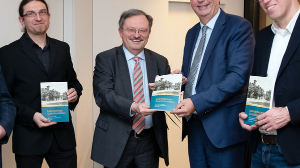 Vier Männer stehen in einem Raum und halten jeweils ein Buch in der Hand. Sie scheinen sich über die Bücher auszutauschen, während ein Mann in der Mitte ein Buch an einen anderen Mann übergibt. Alle wirken lächelnd und freundlich.