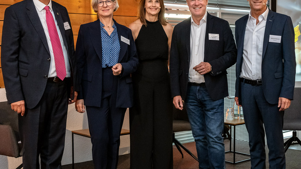 Fünf Personen stehen nebeneinander in einem Raum. Sie lächeln und tragen formelle Kleidung. Im Hintergrund ist ein Bildschirm sichtbar, der eine Einladung zu einer Veranstaltung mit dem Titel "Wirtschaft im Gespräch" anzeigt, datiert auf den 22. Juli 2023.