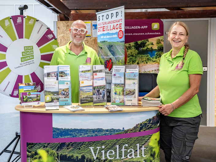 Zwei Personen stehen an einem Informationsstand mit einem bunten Plakat und mehreren Broschüren über lokale Attraktionen. Die Männer trägt eine Brille und hat einen breiten Lächeln. Die Frau hat lange Haare und lächelt ebenfalls. Im Hintergrund ist eine Landschaftsaufnahme zu sehen. (Text KI generiert)