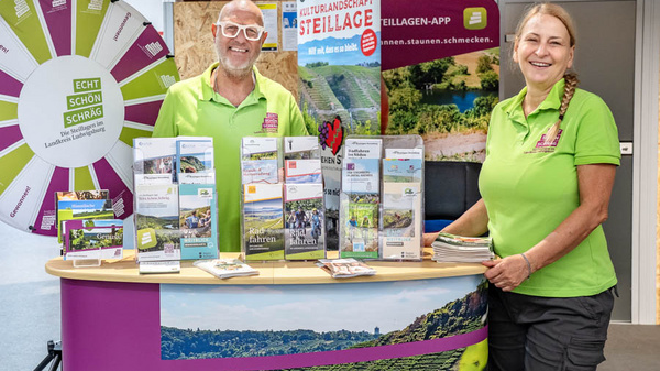 Zwei Personen stehen an einem Informationsstand mit einem bunten Plakat und mehreren Broschüren über lokale Attraktionen. Die Männer trägt eine Brille und hat einen breiten Lächeln. Die Frau hat lange Haare und lächelt ebenfalls. Im Hintergrund ist eine Landschaftsaufnahme zu sehen. (Text KI generiert)