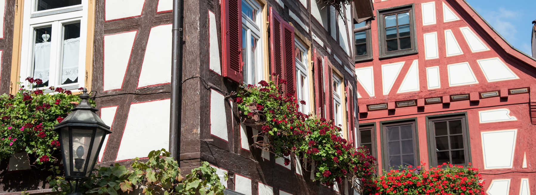 Header Bild: Blick auf zwei historische Fachwerkhäuser mit bunten Blumenkästen. Das linke Haus hat weiße Wände mit dunkelbraunen Balken und rote Fensterläden, während das rechte Haus rote und weiße Wände zeigt. Beide Gebäude sind von Pflanzen umgeben, die eine einladende Atmosphäre schaffen.