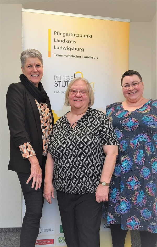 Drei Frauen stehen lächelnd vor einem Banner des Pflegestützpunkts Landkreis Ludwigsburg. Die Frauen tragen unterschiedlich gemusterte Kleidung und scheinen Teil des Teams zu sein. Hintergrund zeigt das Logo und den Namen der Einrichtung.