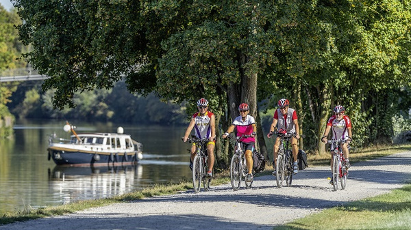 Four cyclists ride along a path beside a calm river, with green trees lining the bank. A small boat glides smoothly on the water, creating a serene outdoor scene. The setting suggests a pleasant day for outdoor activities.
