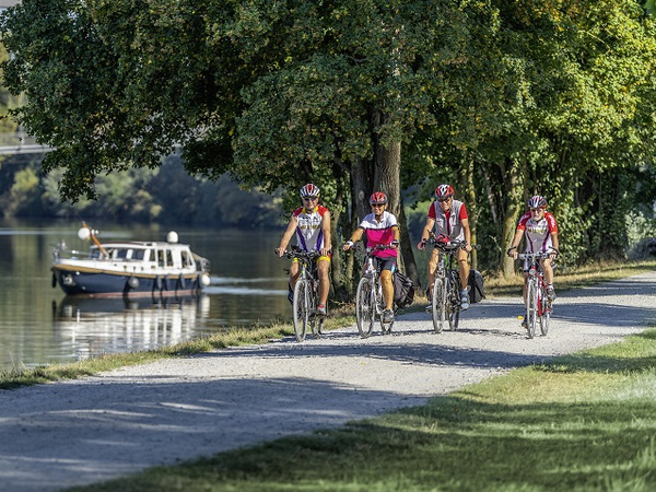 Vier Radfahrer in Sportkleidung fahren entlang eines Weges am Wasser. Im Hintergrund ist ein Boot auf dem Kanal sichtbar, während Bäume das Ufer säumen.