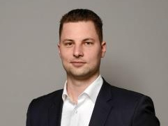 Un uomo in piedi indossa un abito scuro e una camicia bianca. Ha un'espressione seria e si trova di fronte a uno sfondo grigio. La sua mano destra è appoggiata sul fianco mentre l'altra è rilassata lungo il corpo.