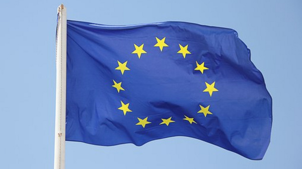A blue flag with a circle of twelve yellow stars arranged in a circle, symbolizing unity. The flag is waving against a clear sky, representing the European Union.