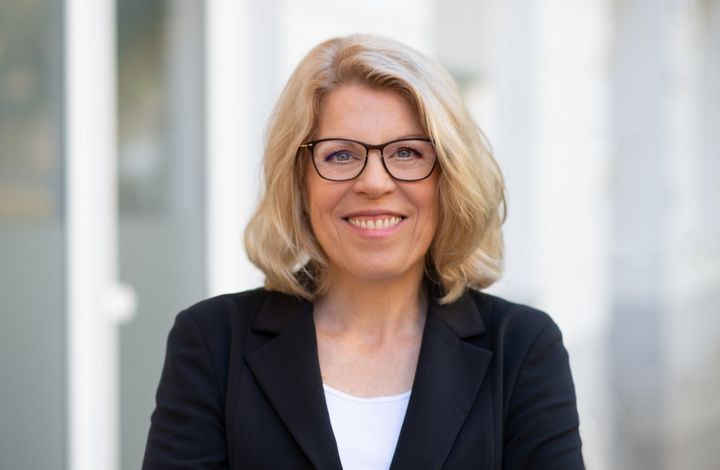 Eine Frau mit blonden, lockigen Haaren und Brille lächelt freundlich in die Kamera. Sie trägt einen schwarzen Blazer und hat die Arme vor der Brust verschränkt. Im Hintergrund sind unscharfe, helle Strukturen zu erkennen.