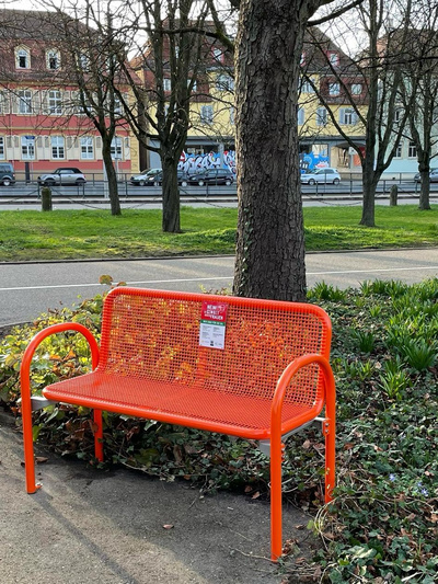 Una panchina arancione si trova in un parco, accanto a un albero e circondata da piante verdi. Sullo schienale della panchina è presente un'etichetta visibile. Sullo sfondo si intravedono edifici e una strada con delle auto.