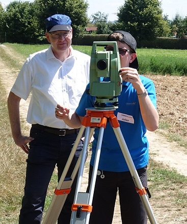 Zwei Personen stehen im Freien auf einem Weg. Eine Person in blauer T-Shirt und schwarzer Kappe bedient ein Theodoliten-Stativ, während die andere Person in weißem Hemd zuschaut. Im Hintergrund sind grüne Felder zu sehen.