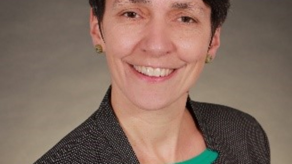 Portrait d'une femme souriante, portant un blazer à motifs et un haut vert. Elle a les cheveux courts et foncés. L'arrière-plan est neutre, mettant en valeur son expression amicale.