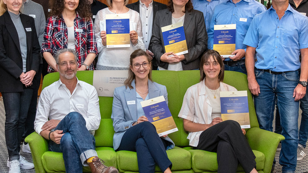 Eine Gruppe von 17 Personen steht und sitzt auf einem grünen Sofa in einem Raum mit Holzverkleidung. Die Personen halten Urkunden in den Händen und lächeln. Es handelt sich um eine Feier oder Zeremonie zur Auszeichnung.